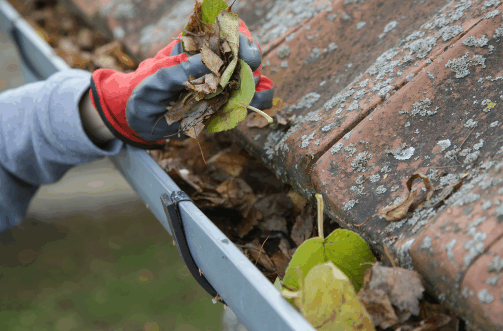 Gloved hand clearing leaves and debris from a home gutter to ensure proper water flow