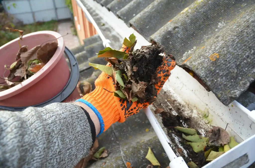 Gloved hand clearing leaves and debris from a home gutter to ensure proper water flow